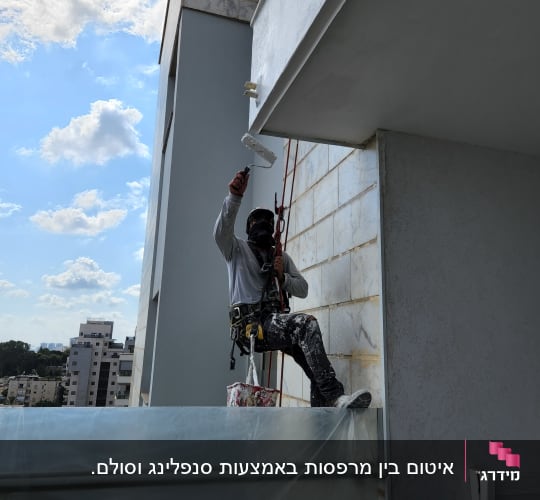 אדם צובע קיר חיצוני עם רולר על חבל